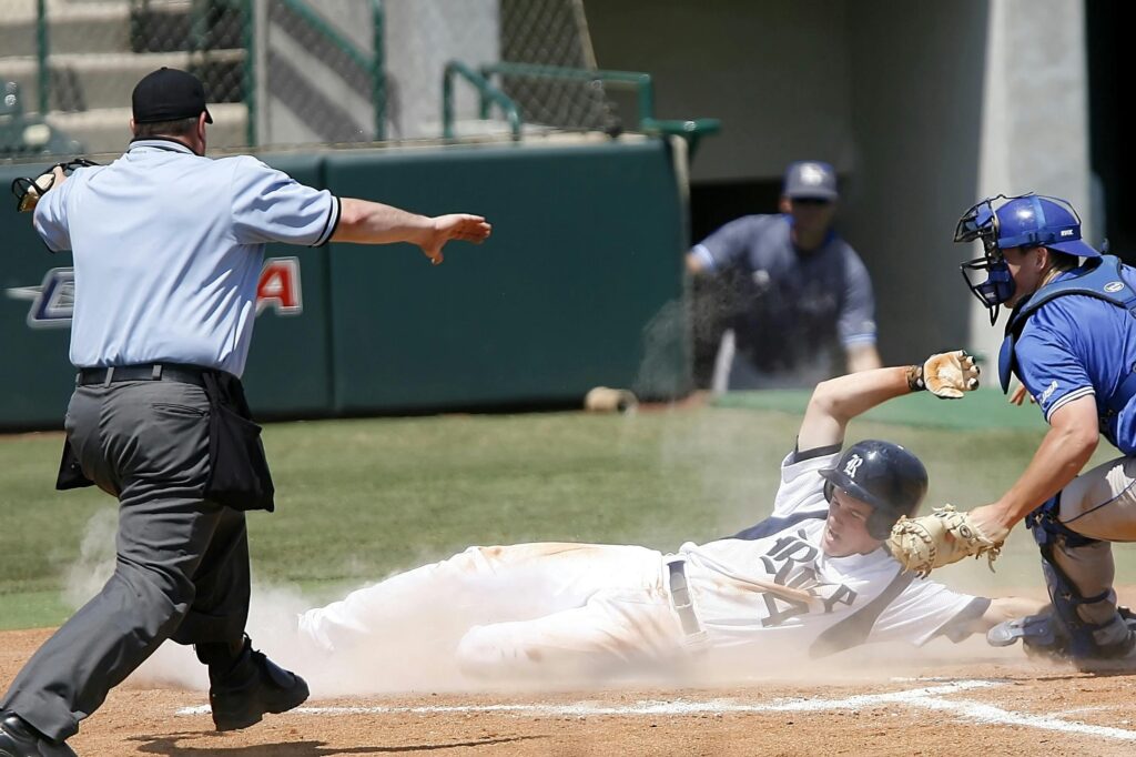 A baseball player in a white uniform slides into home plate through a cloud of dust as the catcher in blue tries to make the tag and the umpire watches closely.
