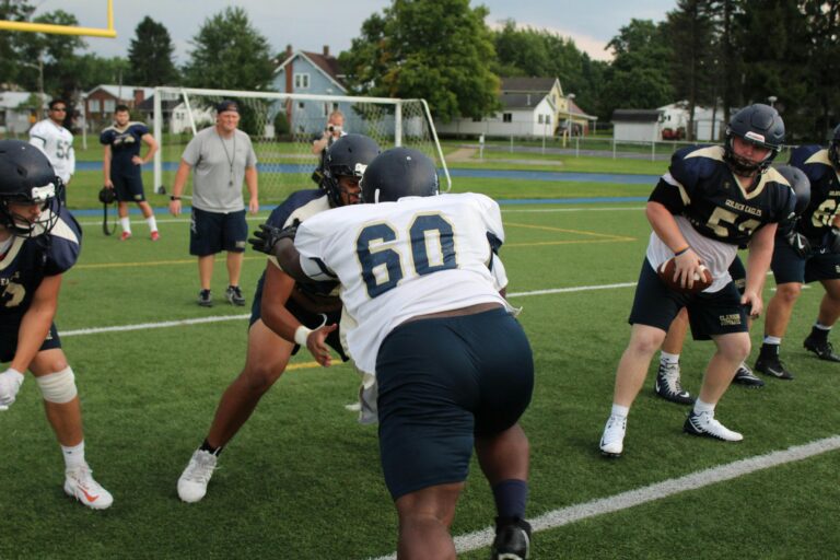 American football players in "Golden Eagles" uniforms engage in a practice drill on a green field with coaches watching in the background.