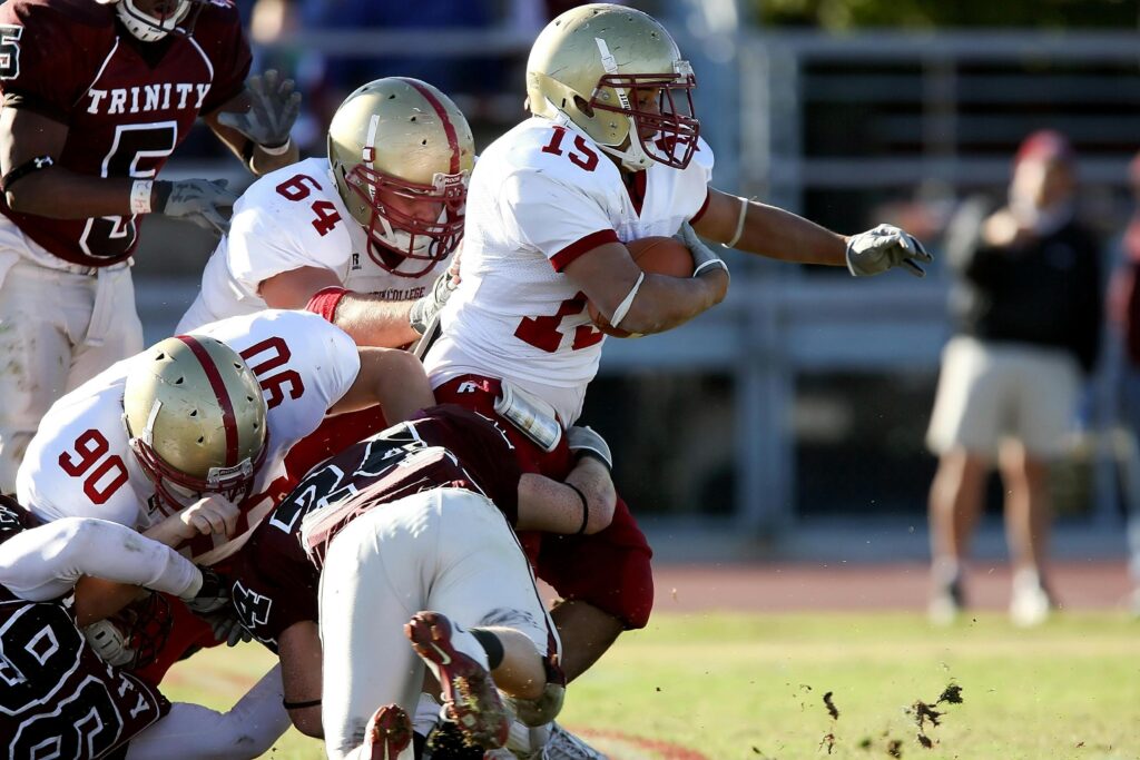 A football player in a white and gold uniform sprints with the ball while being tackled by several opposing players in maroon jerseys.