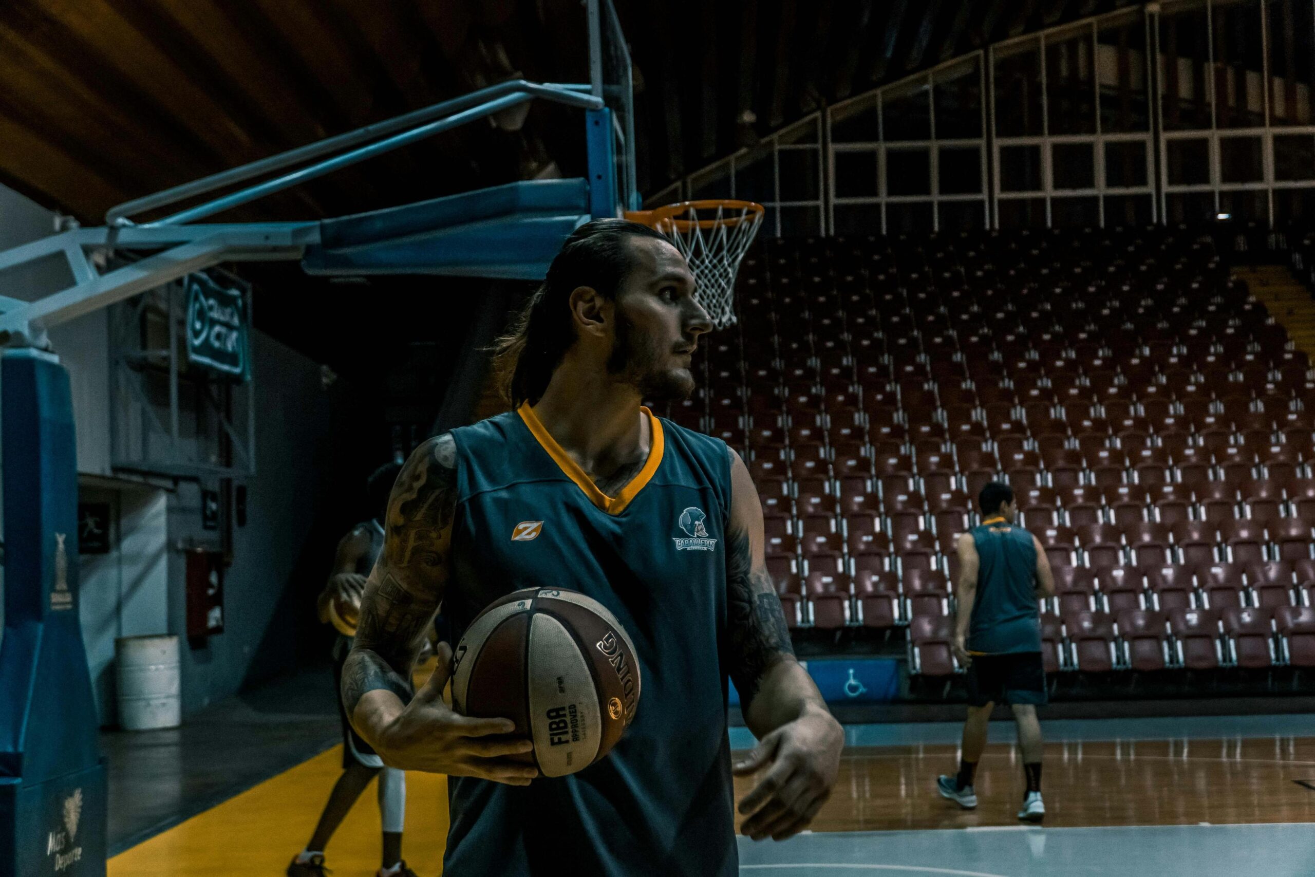 A basketball player with tattoos holds a ball on an indoor court in front of rows of empty stadium seating.