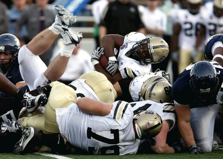 A group of American football players in white and gold uniforms are piled together in a tackle on the field.