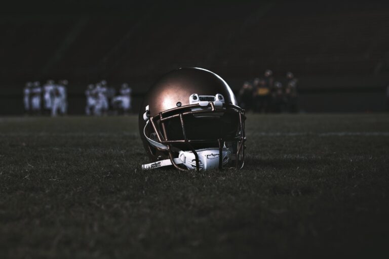 A bronze-colored American football helmet sits on a dark grass field with players blurred in the background.