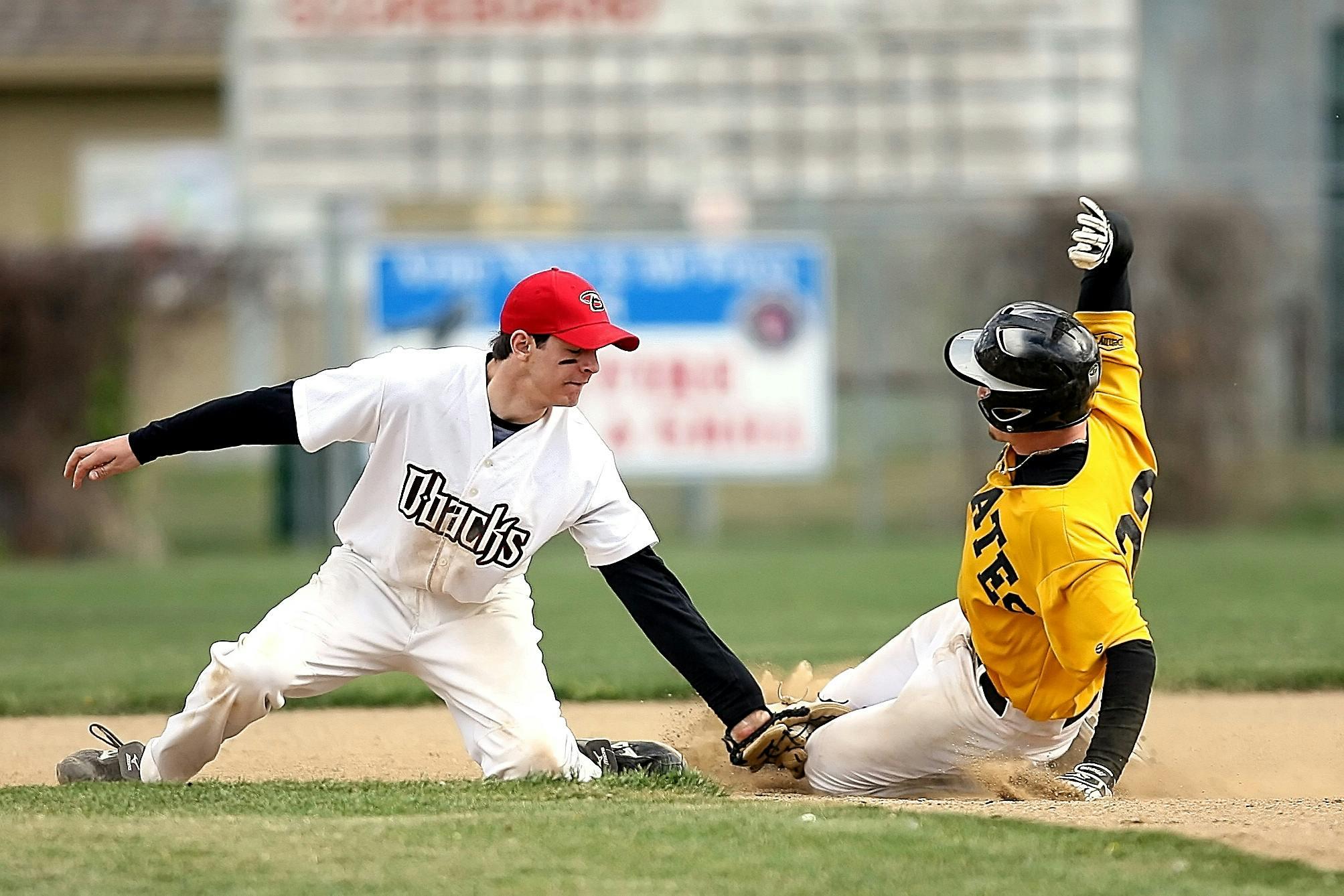 A baseball fielder in a white uniform reaches to tag a runner in a yellow jersey sliding headfirst into a base.
