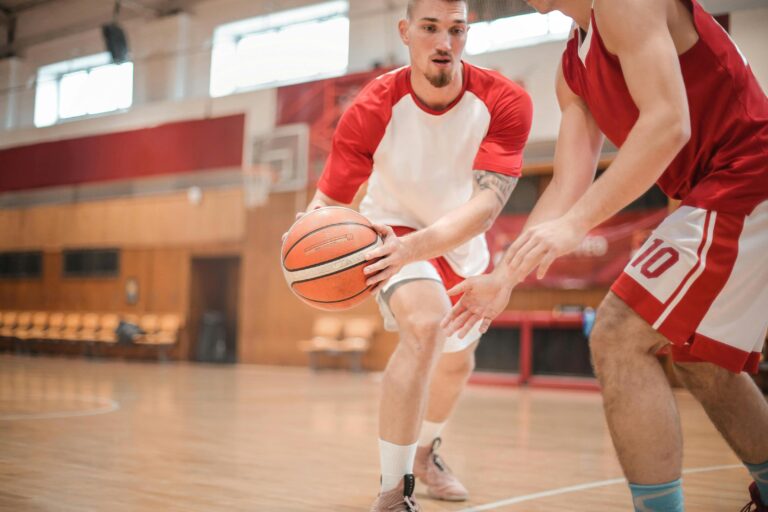 A basketball player in a red and white jersey dribbles the ball while being closely guarded by an opponent during an indoor game.