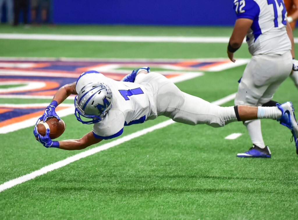 A football player in a white and blue uniform with the number 1 dives horizontally through the air to catch a football just above the green turf.