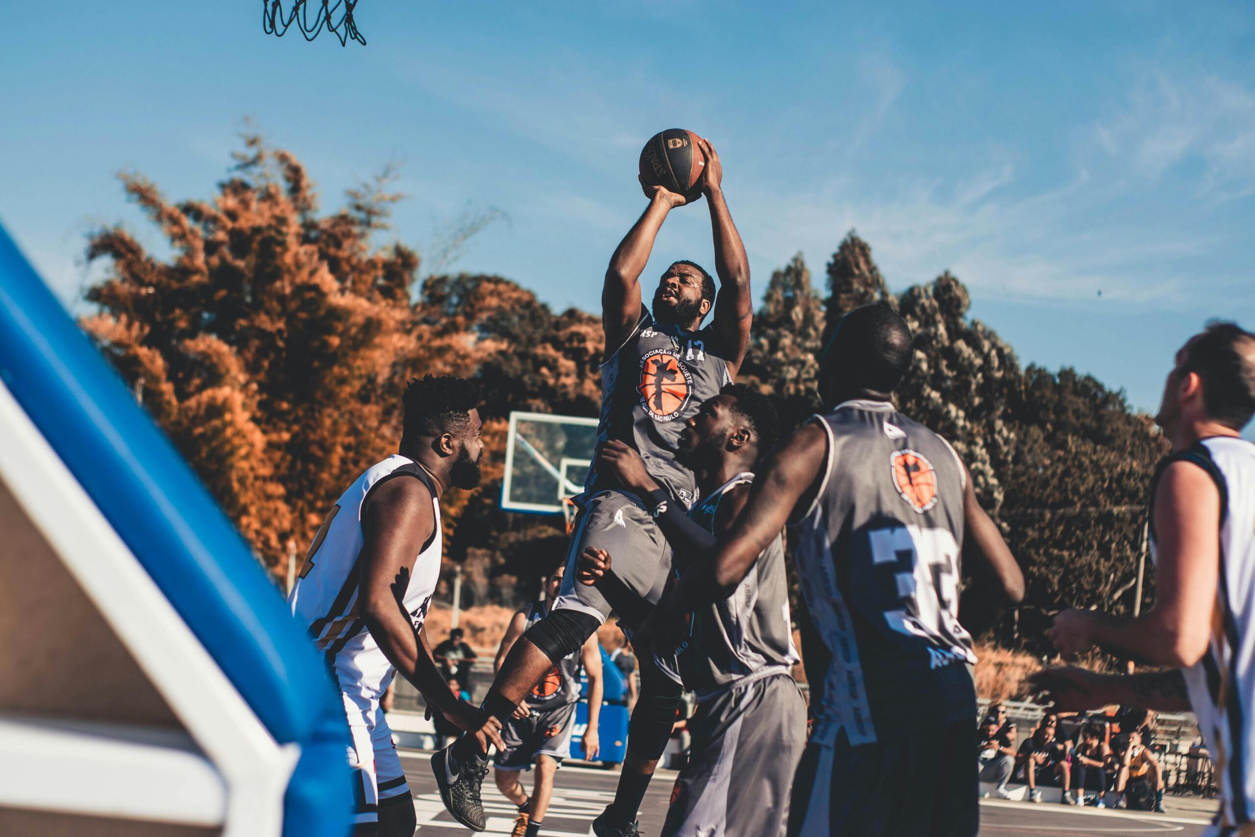 A basketball player in a grey jersey jumps high to shoot the ball during an outdoor game surrounded by other players.