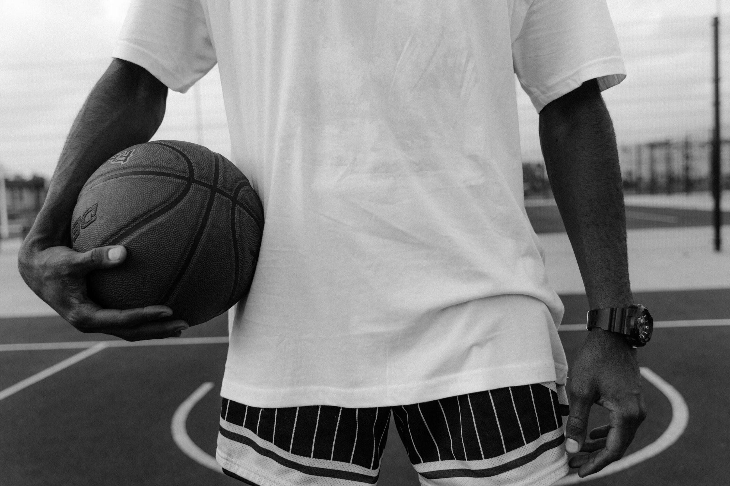 A black and white, mid-section shot of a person standing on an outdoor basketball court holding a basketball under one arm.