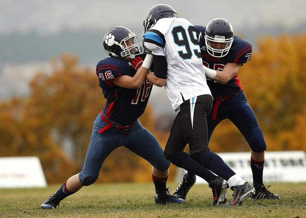 Three football players in full uniform engage in a physical block on a grass field during a game.