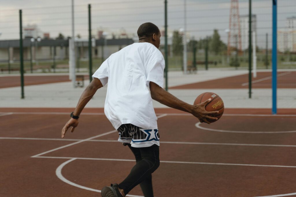 A basketball player from behind, wearing a white t-shirt and striped shorts, dribbles a ball on an outdoor court.