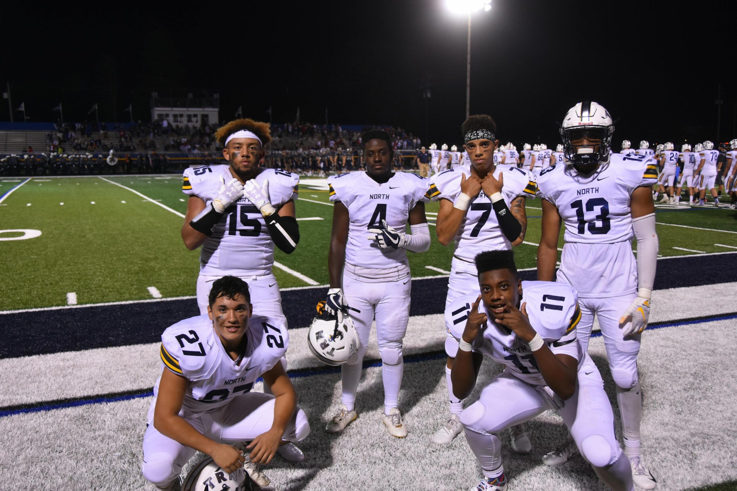 Six football players in white uniforms with yellow and black accents pose together on a field at night.