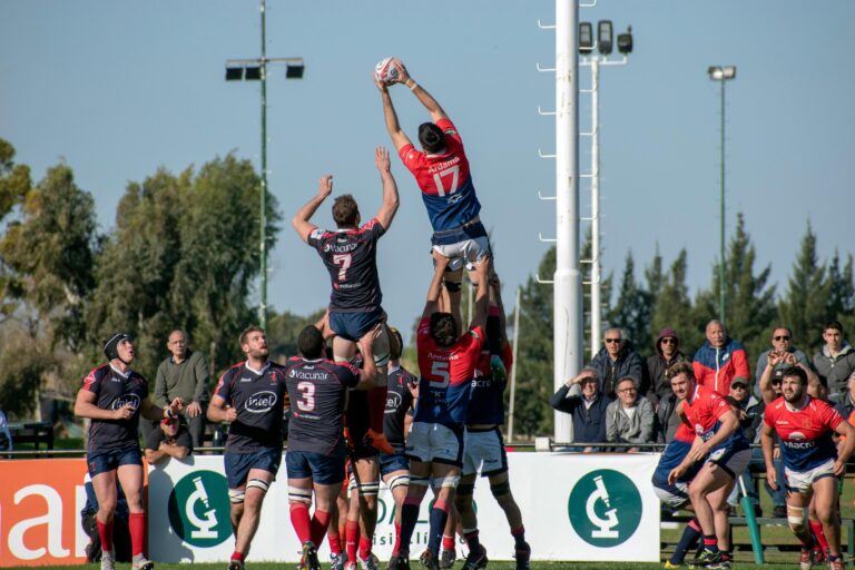 Rugby players in red and blue jerseys perform a line-out, lifting a teammate high into the air to catch the ball.