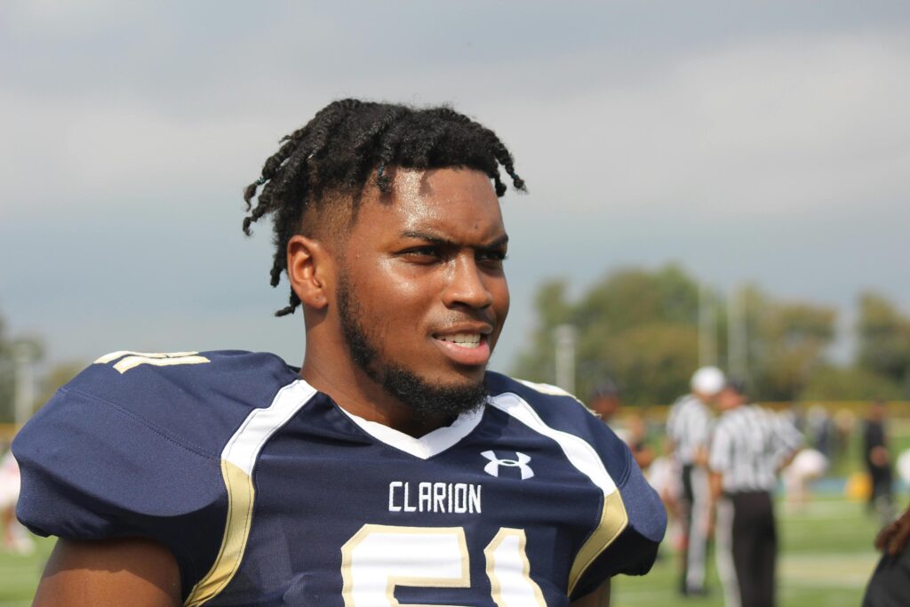 A close-up portrait of an American football player in a navy blue and gold Clarion jersey on a sunny field.