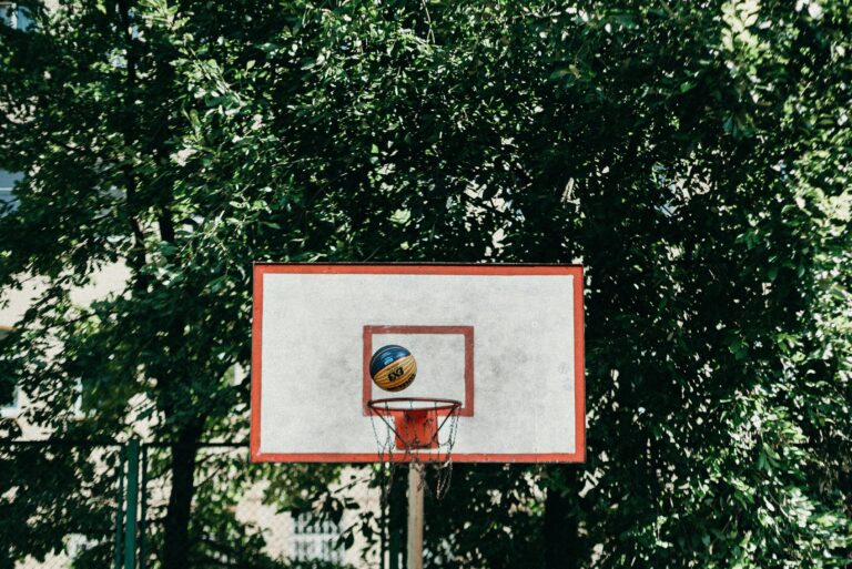 A basketball bounces off a weathered backboard against a backdrop of dense green trees.