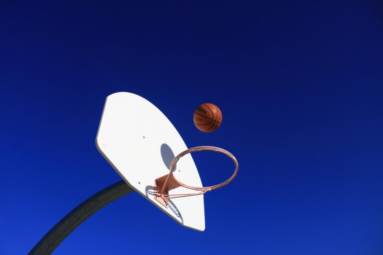 A low-angle shot of a basketball suspended in the air just above a white backboard and rim against a clear, deep blue sky.
