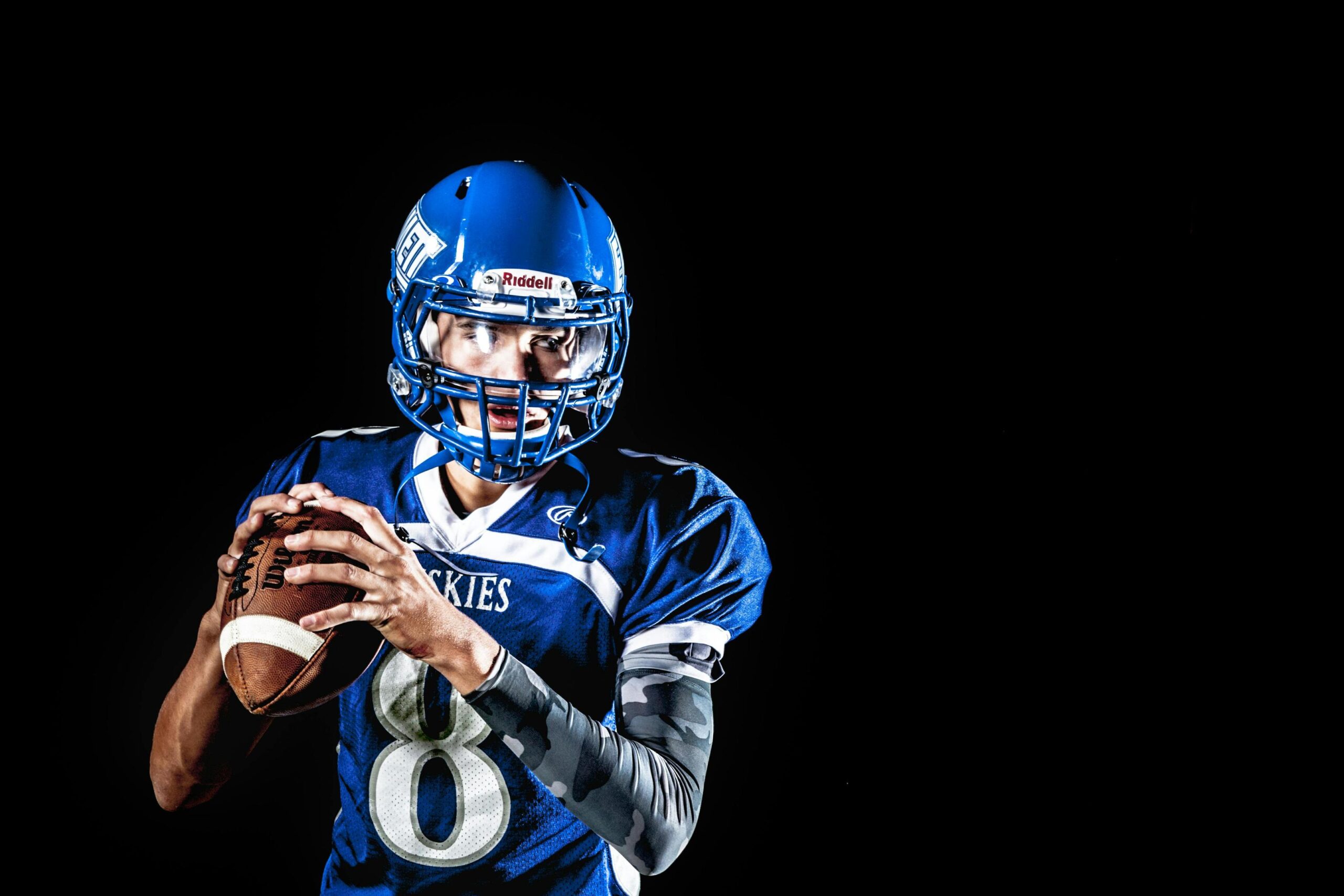 A football player in a blue Huskies jersey and helmet looks to the side while holding a football against a black background.