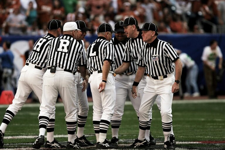 A group of American football officials in traditional black-and-white striped jerseys and white pants huddle together on the field during a game.