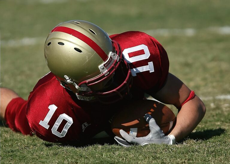 An American football player in a red and gold uniform lies on the grass while firmly clutching a football.