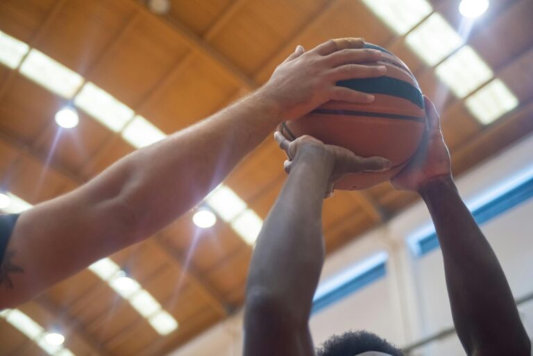 A basketball player with tattoos holds a ball on an indoor court in front of rows of empty stadium seating.