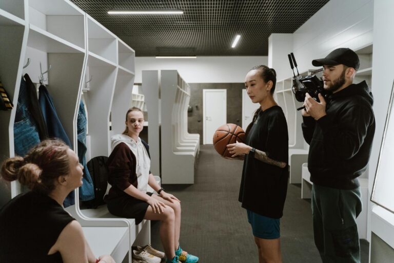Inside a locker room, a videographer films a group of athletes as one holds a basketball and speaks.