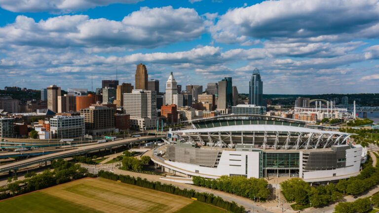 An aerial view of a large modern sports stadium situated within a bustling city skyline under a cloudy blue sky.