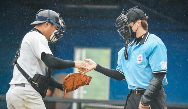 A baseball umpire in a light blue shirt hands a ball to a catcher in a white jersey and full protective gear during a light rain.