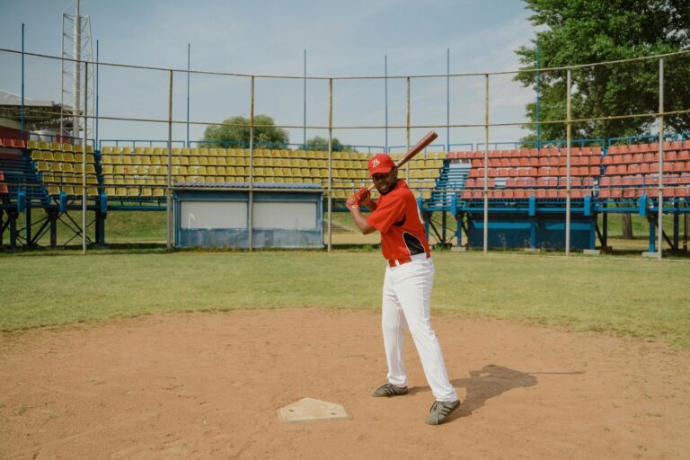 A baseball player in a red jersey and white pants stands at home plate, holding a bat and ready to swing.