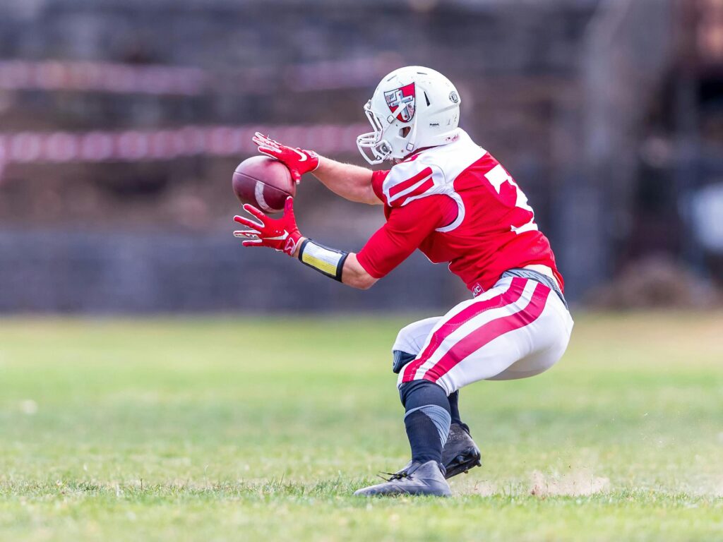 A football player in a red and white uniform reaches out to catch a football during a game on a grass field.