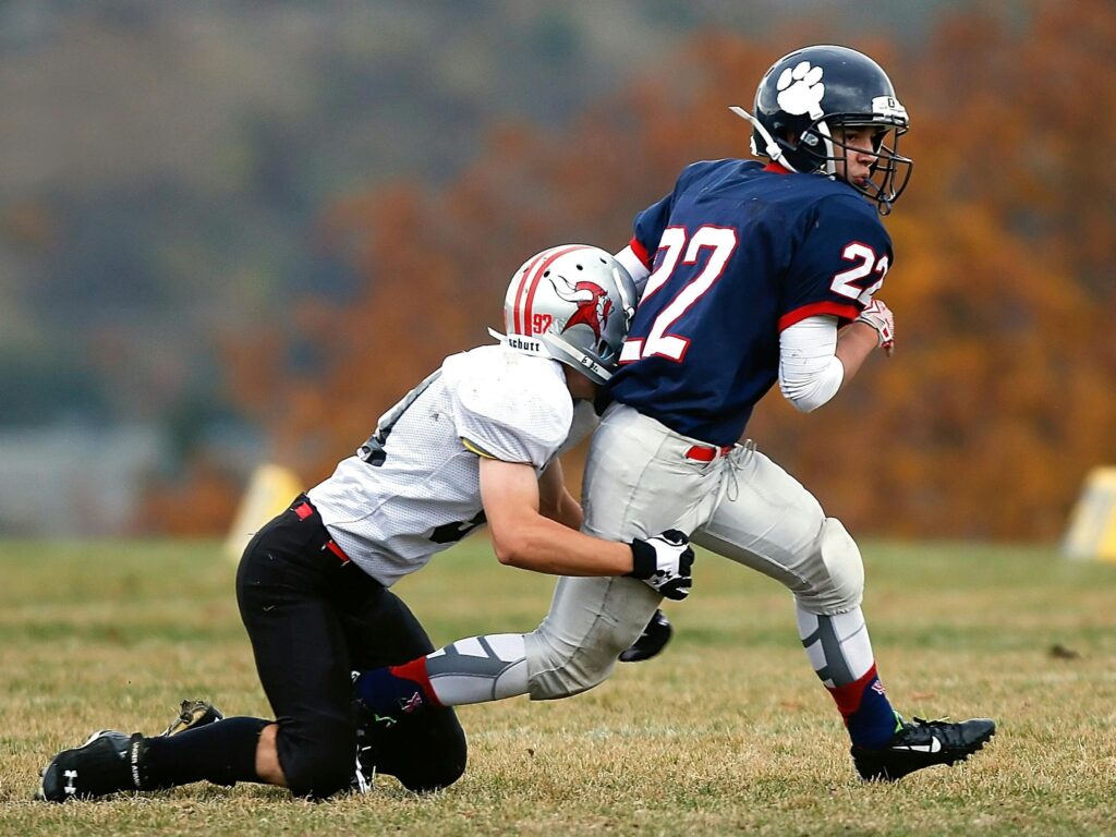 An action shot of a football player in a white jersey being tackled from behind by a player in a dark blue jersey with the number 22.
