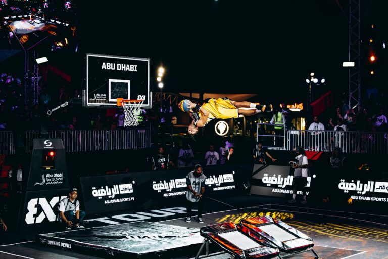 An acrobatic basketball player in a yellow uniform performs a mid-air backflip while holding a basketball near a hoop during a dunk show in Abu Dhabi.