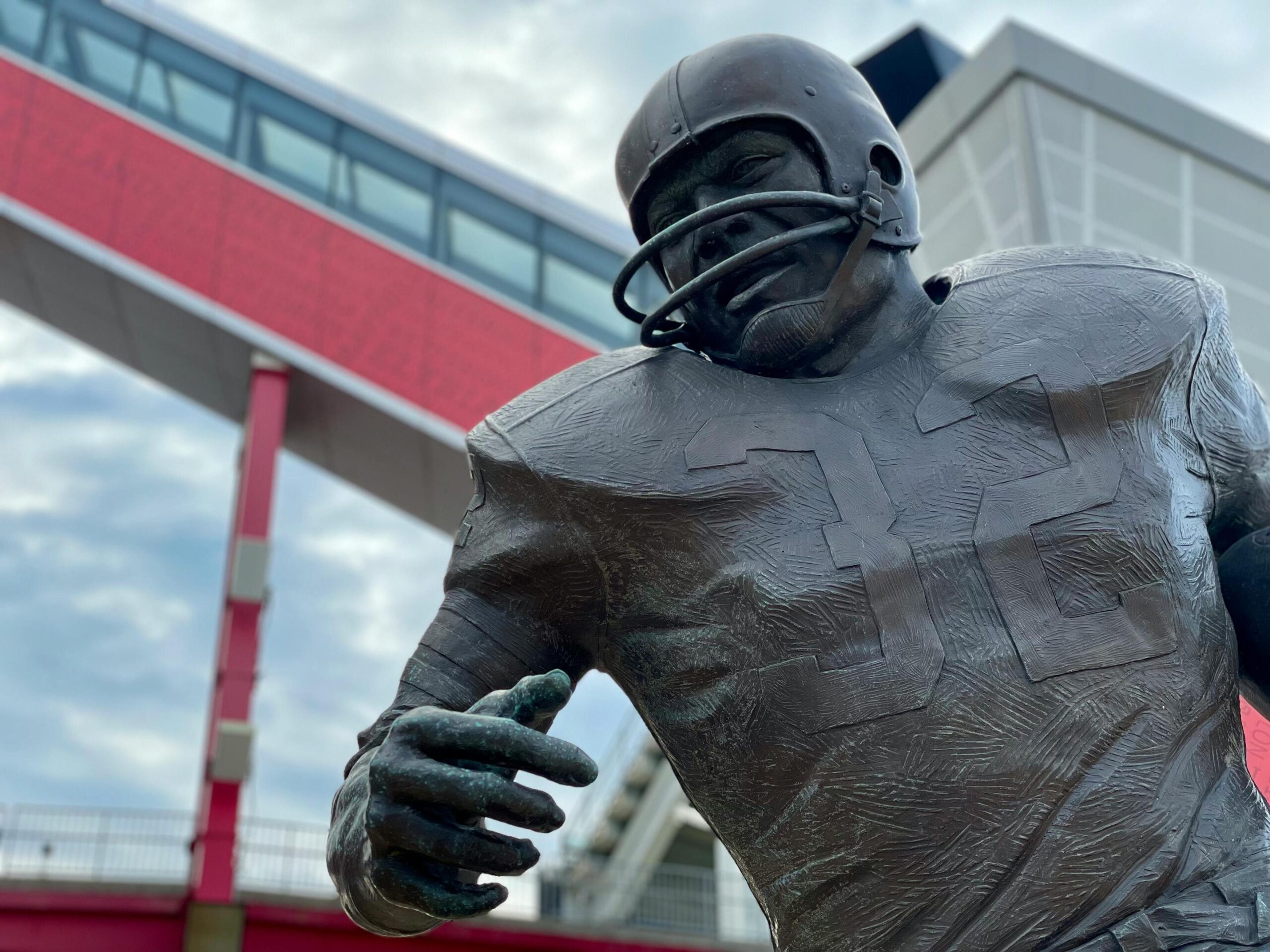 A bronze statue of an American football player wearing a vintage helmet and jersey stands against a cloudy sky.