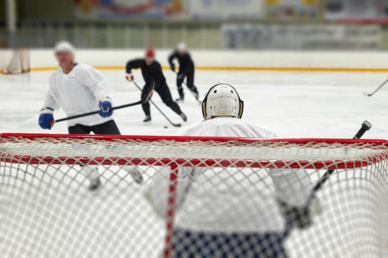 A view from behind a hockey goalie in a white jersey looking out toward players on the ice rink.
