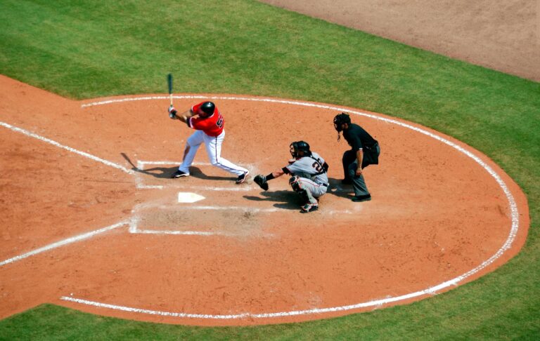 A high-angle shot of a baseball game showing a batter in a red jersey swinging at a pitch, with a catcher and umpire positioned behind home plate.