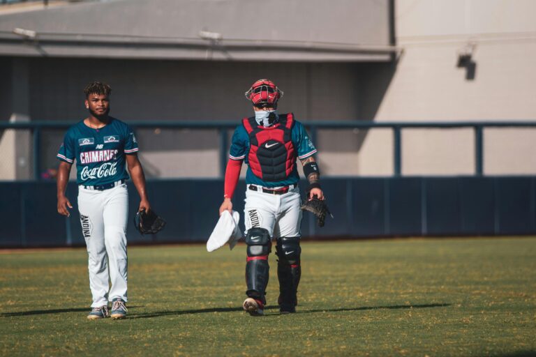 Two baseball players in teal and white uniforms walk across a grassy field, with one player wearing full catcher's gear and carrying a white towel.