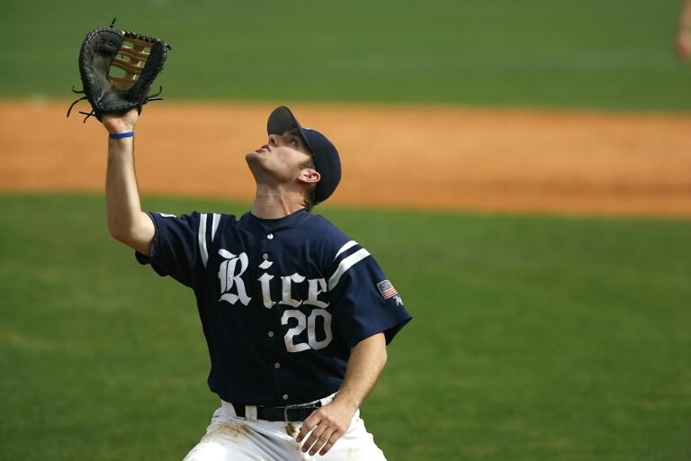 A baseball player in a navy blue "Rice" jersey looks upward while holding his glove high to catch a fly ball.